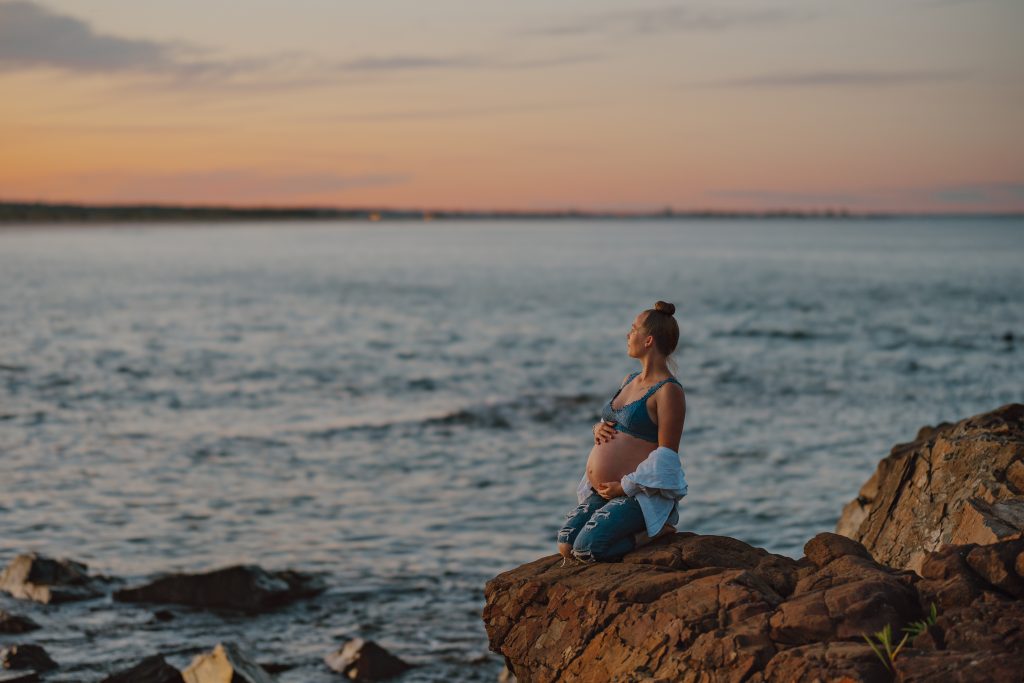 maternity photo of pregnant woman sitting on a rocky outcropping at the ocean