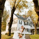 Bride and groom kiss outside a historic white New England home during their fall wedding, surrounded by autumn foliage and fallen leaves.