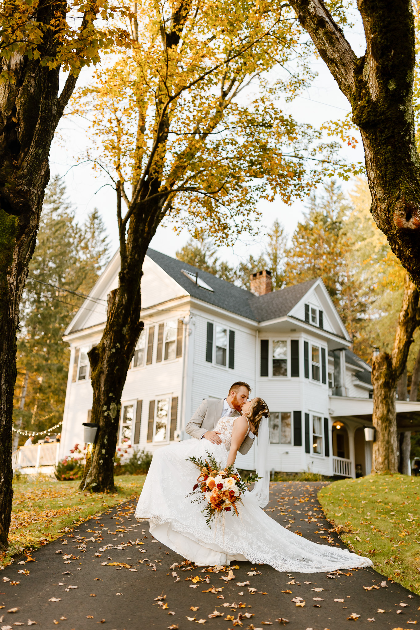 Bride and groom kiss outside a historic white New England home during their fall wedding, surrounded by autumn foliage and fallen leaves.