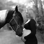 Pregnant woman in a black maternity dress standing beside her horse during an outdoor maternity session in New Hampshire's Lakes Region.