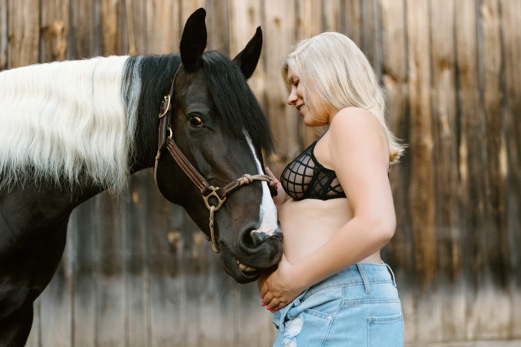 Pregnant woman in a black maternity dress standing beside her horse during an outdoor maternity session in New Hampshire's Lakes Region.